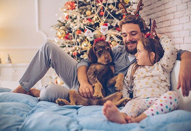 Padre y su adorable hija recostados en la cama con su perro. Al fondo, árbol de Navidad.