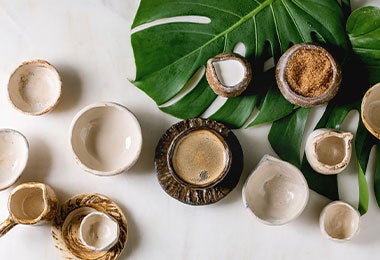 Taza de cerámica con café expreso servido con azúcar de caña, jarra de crema y muchos cuencos de cerámica vacíos sobre una hoja de monstera sobre un fondo de mármol blanco.