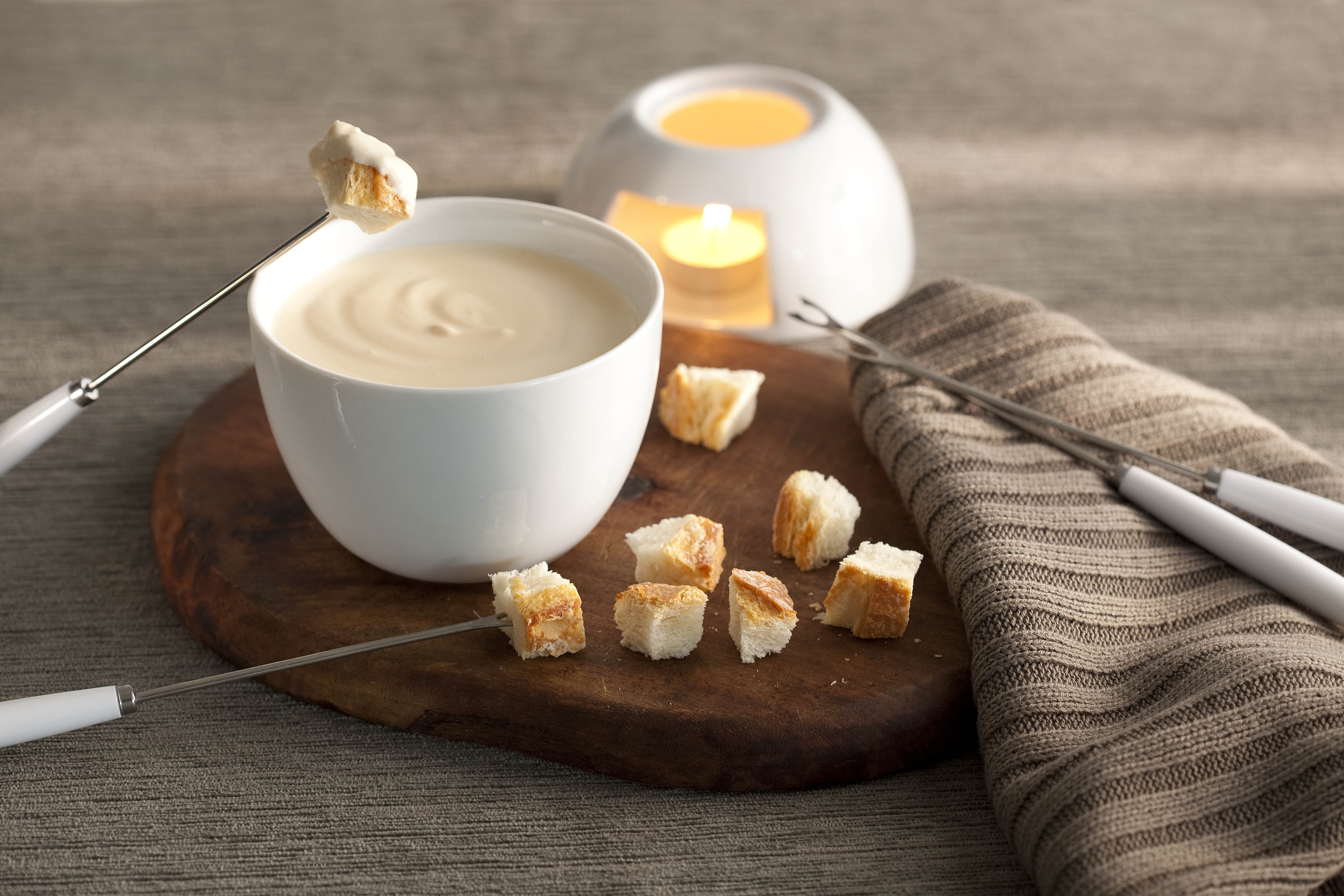 fotografía en tonos grises, marrones y banco visto de frente. Contiene una tabla de madera redonda con un recipiente blanco redondo que contiene la fondue, 4 tenedores de fondue y trozos de pan alrededor. fotografía en tonos grises, marrones y banco visto de frente. Contiene una tabla de madera redonda con un recipiente blanco redondo que contiene la fondue, 4 tenedores de fondue y trozos de pan alrededor.