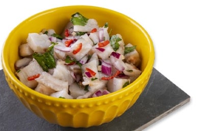 Ceviche con leche de tigre, cebolla morada, cilantro y limón en un recipiente amarillo sobre un fondo blanco