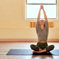 Mujer practicando yoga en casa encima de una esterilla y con los brazos estirados hacia arriba