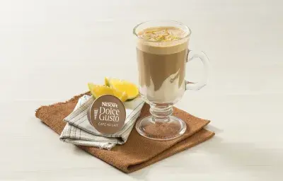 fotografía en tonos blanco y marrón tomada de frente, contiene un paño marrón con un vaso transparente con la bebida de NESCAFÉ Dolce Gusto Café Au Lait con Crema de Leche NESTLÉ. Al lado hay una cápsula de Dolce Gusto y al fondo rodajas de naranja fotografía en tonos blanco y marrón tomada de frente, contiene un paño marrón con un vaso transparente con la bebida de NESCAFÉ Dolce Gusto Café Au Lait con Crema de Leche NESTLÉ. Al lado hay una cápsula de Dolce Gusto y al fondo rodajas de naranja