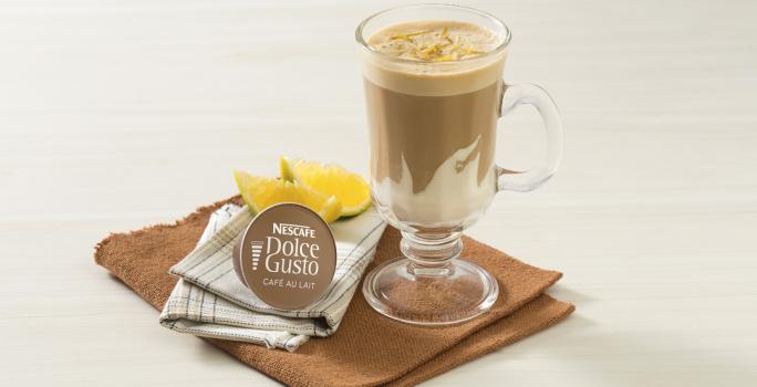 fotografía en tonos blanco y marrón tomada de frente, contiene un paño marrón con un vaso transparente con la bebida de NESCAFÉ Dolce Gusto Café Au Lait con Crema de Leche NESTLÉ. Al lado hay una cápsula de Dolce Gusto y al fondo rodajas de naranja